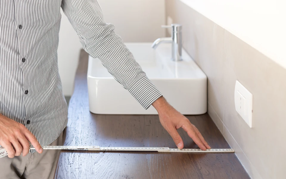 Une personne en chemise rayée mesure avec une règle métallique graduée l'espace entre un lavabo moderne blanc et un mur dans une salle de bain contemporaine aux tons beiges